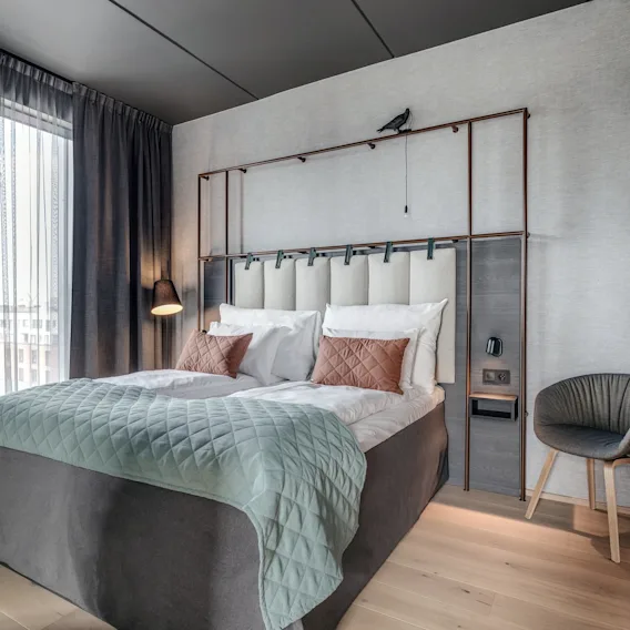En seng med hvite og brune puter står mot et polstret hodegjerde. En svart lampe henger i et moderne, lyst rom med tregulv og et vindu med mørke gardiner.