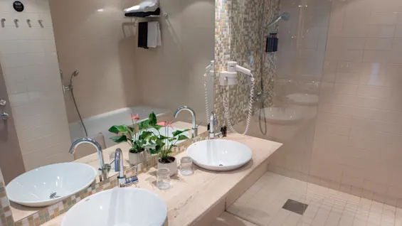Två vita handfat står på en marmorbänk med en spegel ovanför, omgiven av beige kakelväggar. En liten krukväxt och toalettartiklar pryder utrymmet. En dusch och handdukar syns.