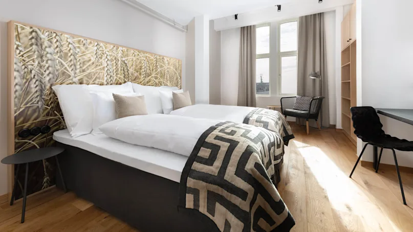 A neatly made bed with patterned throw blankets in a room with modern furnishings and a large window, exuding a cozy atmosphere.