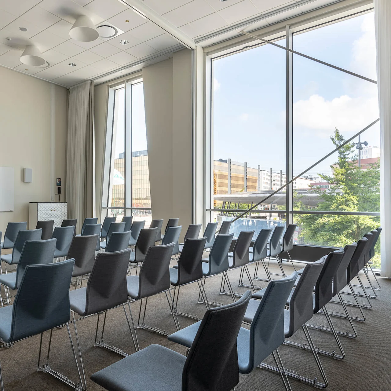 Rows of empty chairs are arranged in a conference room, facing a whiteboard on the front wall. Large windows reveal a view of trees and buildings outside.
