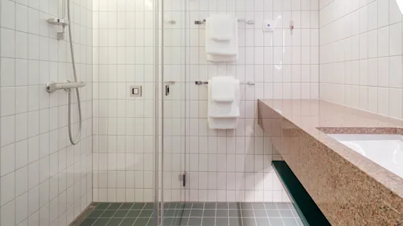 En dusch med handmunstycke och slang, sitter mot vitkaklade väggar. Två vita handdukar hänger på stänger. Bredvid finns en stenskiva med integrerad diskho.