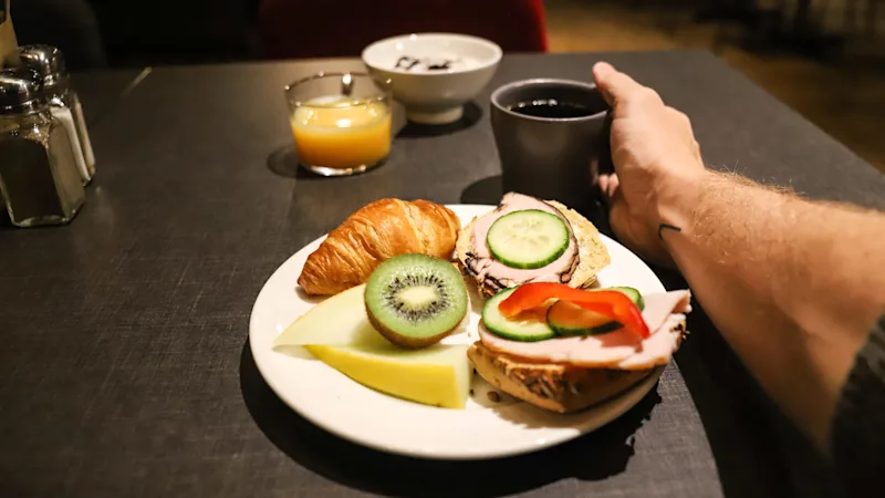 A breakfast plate featuring a croissant, slices of cheese, kiwi, and sandwiches topped with cold cuts, cucumber, and bell pepper. A hand is holding a cup of black coffee.