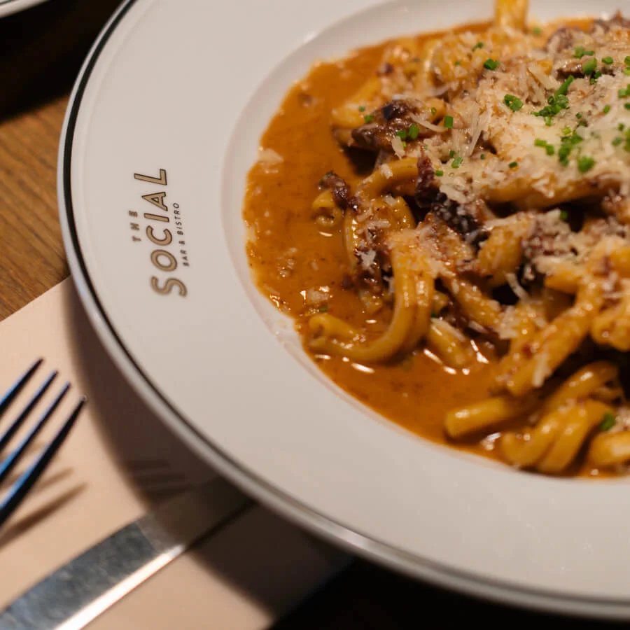 En tallrik med krämig pasta med sås och örter på ett träbord. I närheten finns bestick och en servett. På tallriken står "THE SOCIAL BAR & BISTRO."