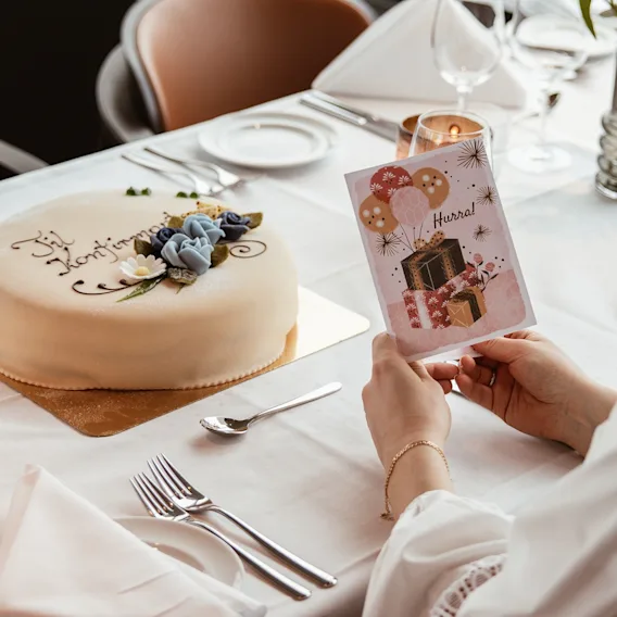 En person leser et gratulasjonskort med 'Hurra' ved et dekket bord med en dekorert kake, som indikerer en feiring. 🎈