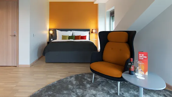 Ett modernt hotellrum med en prydligt bäddad säng med färgglada kuddar mot en orange vägg. En orange stol och ett litet bord med en vinflaska och glas. Text på kort: "GET ME SOMETHING TO EAT"