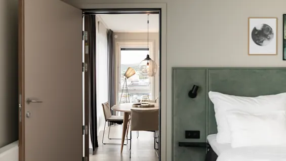 Viihtyisä makuuhuonenäkymä, joka avautuu valoisaan oleskelutilaan ruokapöydällä ja ikkunalla, josta on näkymä kaupunkiin.