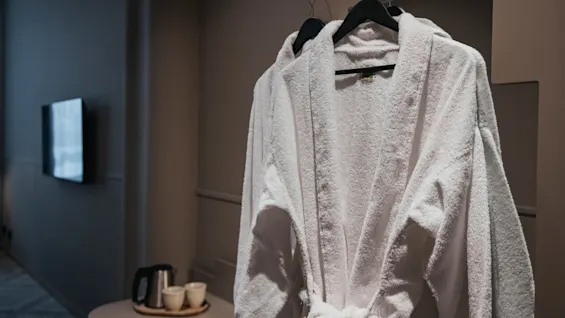 Två mjuka vita badrockar hänger i ett mysigt hotellrum, redo för en avkopplande vistelse. En vattenkokare och koppar står på ett närliggande bord.