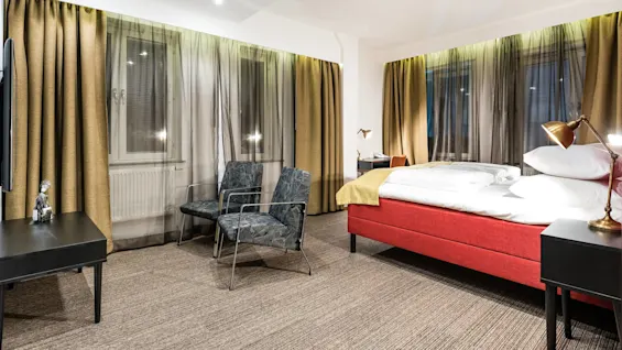 Et moderne hotellrom med en rød seng, to mønstrede stoler og messinglamper, omgitt av brune gardiner og store vinduer, som skaper en koselig atmosfære.