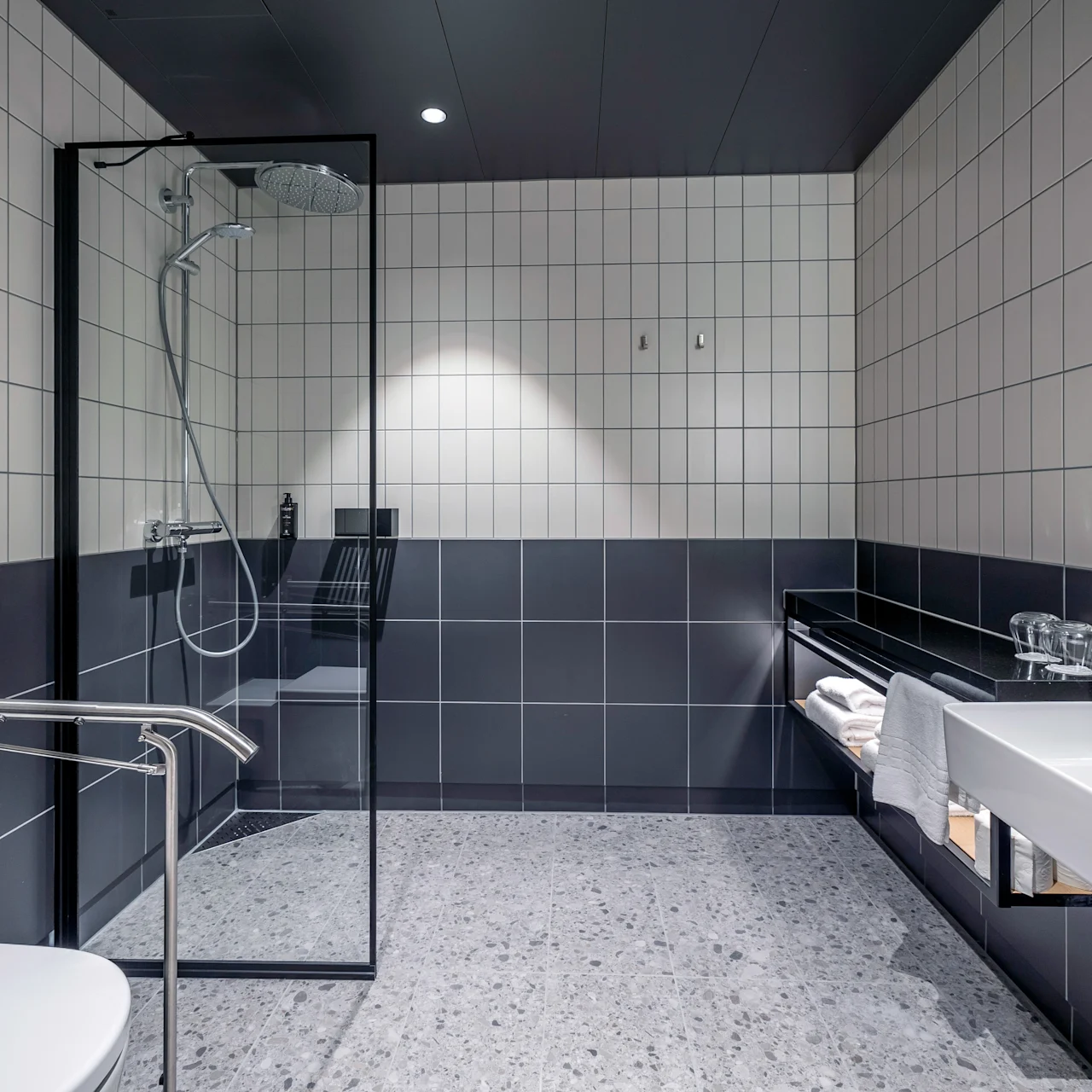 Moderne badeværelse med hvide og grå fliser. En glasinddækket bruser står ved siden af et toilet og en vask med bordplade. Håndklæder er pænt arrangeret på en hylde.