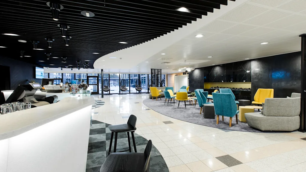 Valoisa, moderni lounge, jossa värikkäät keltaiset ja siniset tuolit matalien pöytien ympärillä matolla, vierekkäin tyylikkään valkoisen baaritiskin kanssa, tumman katon alla.