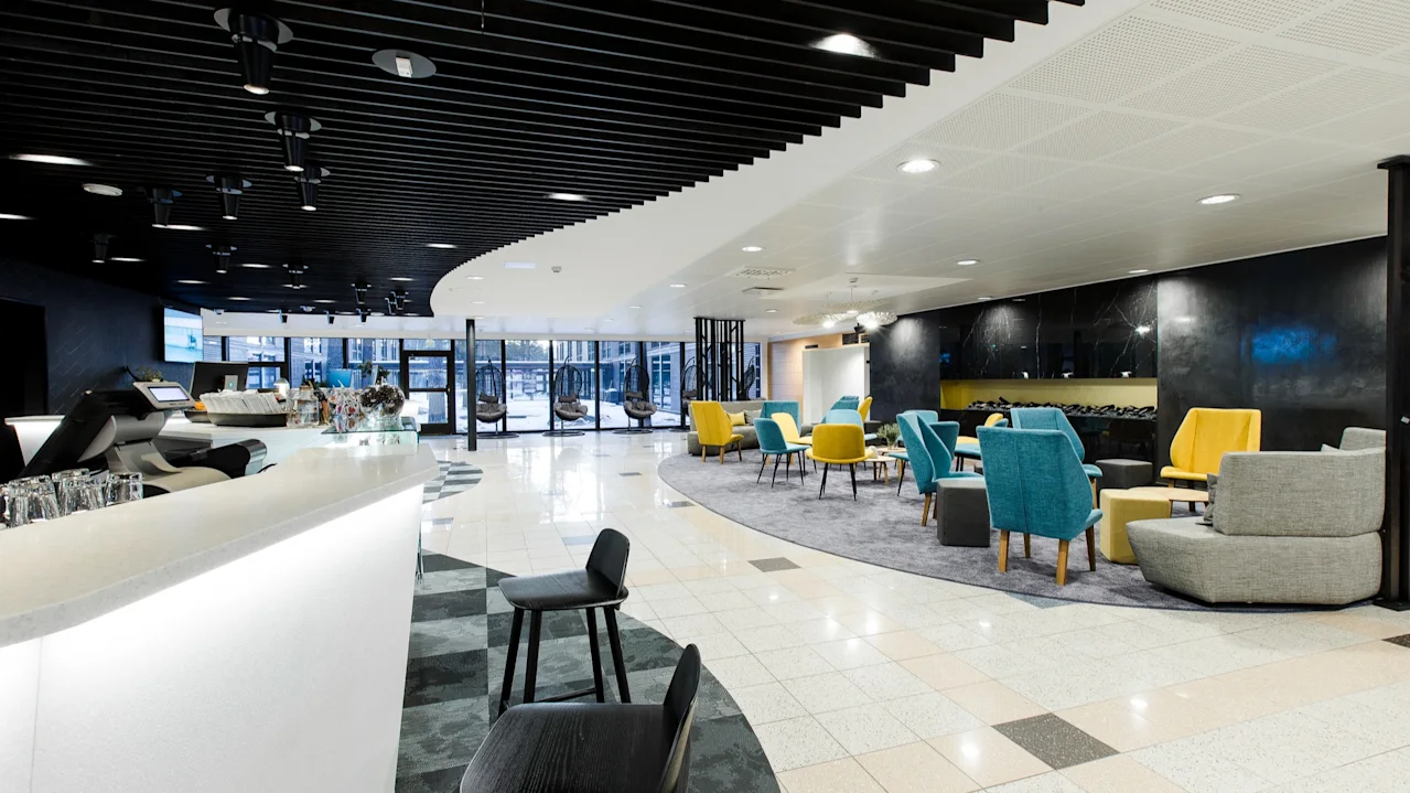 Valoisa, moderni lounge, jossa värikkäät keltaiset ja siniset tuolit matalien pöytien ympärillä matolla, vierekkäin tyylikkään valkoisen baaritiskin kanssa, tumman katon alla.