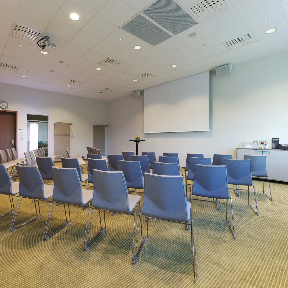Rows of empty gray chairs face a projection screen in a meeting room with beige carpet and a ceiling-mounted projector. A table with refreshments sits on the right.