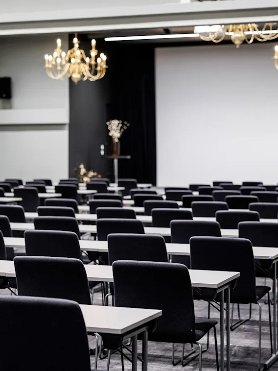 En stor konferansesal satt opp i klasseromsoppsett med rader av sorte stoler, lange bord, lysekroner og et lerret i front.