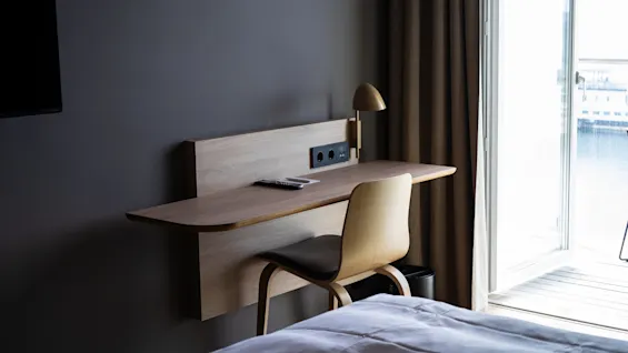 Vegghengt skrivebord med en lampe og fjernkontroll på toppen. En stol står under. Kontekst: ved et vindu med beige gardiner og delvis utsikt over en seng og åpen balkongdør.