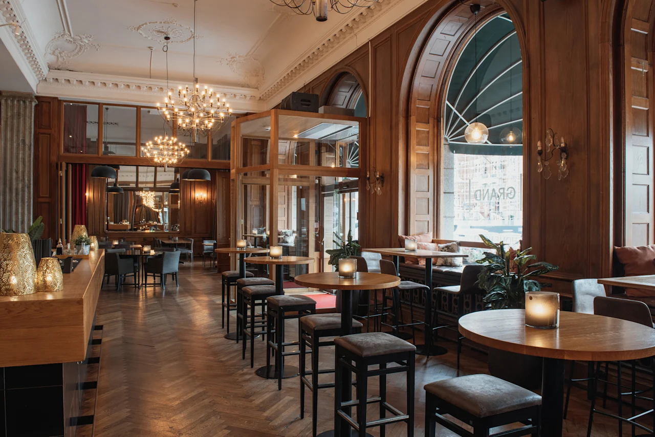 En varm och inbjudande hotellobbybar med träinredning, bekväma sittplatser och stora välvda fönster. GRAND är synligt.