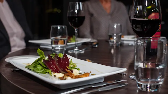 A white rectangular plate with a salad of beets, greens, and cheese sits on a wooden table. Red wine glasses and water glasses are arranged around the dining setting.