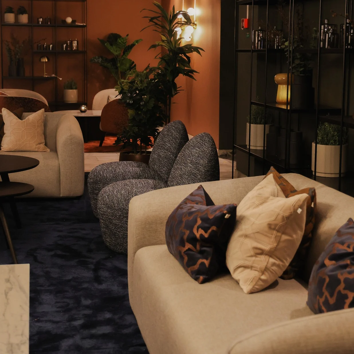 A beige sofa with patterned pillows sits on a dark blue carpet in a cozy, plant-adorned living room. A small round table and decorative items complete the modern setting.