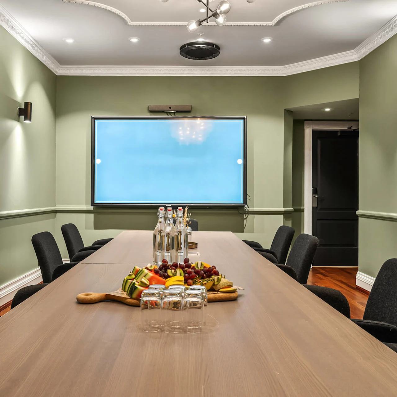 Et langt træbord har frugt, glas og flasker i et grønmalet konferencerum. Sorte stole omgiver bordet, under et loft med spotlights og en central dekorativ armatur.