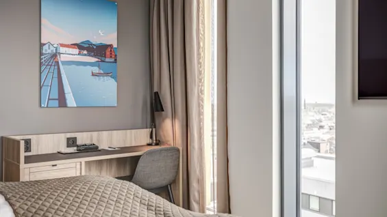 A desk with a lamp and chair sits beside a large window with city views. A colorful seaside painting hangs above. Neutral curtains frame the scene.