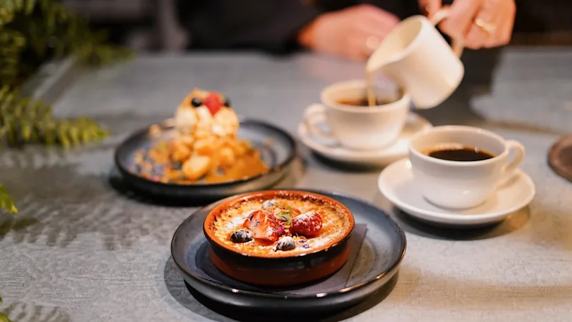 A dessert topped with berries sits on a table beside two cups of coffee. A hand pours cream into one cup, surrounded by a plant.