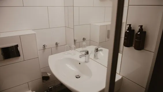 A white ceramic sink with metal faucets is installed on a tiled wall, accompanied by two clear glasses and two soap dispensers. The surrounding environment is a modern bathroom.