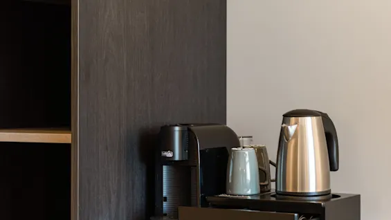 En kaffemaskine, elkedel og kopper på en mørk disk ved siden af et træskab, klar til en varm drik.