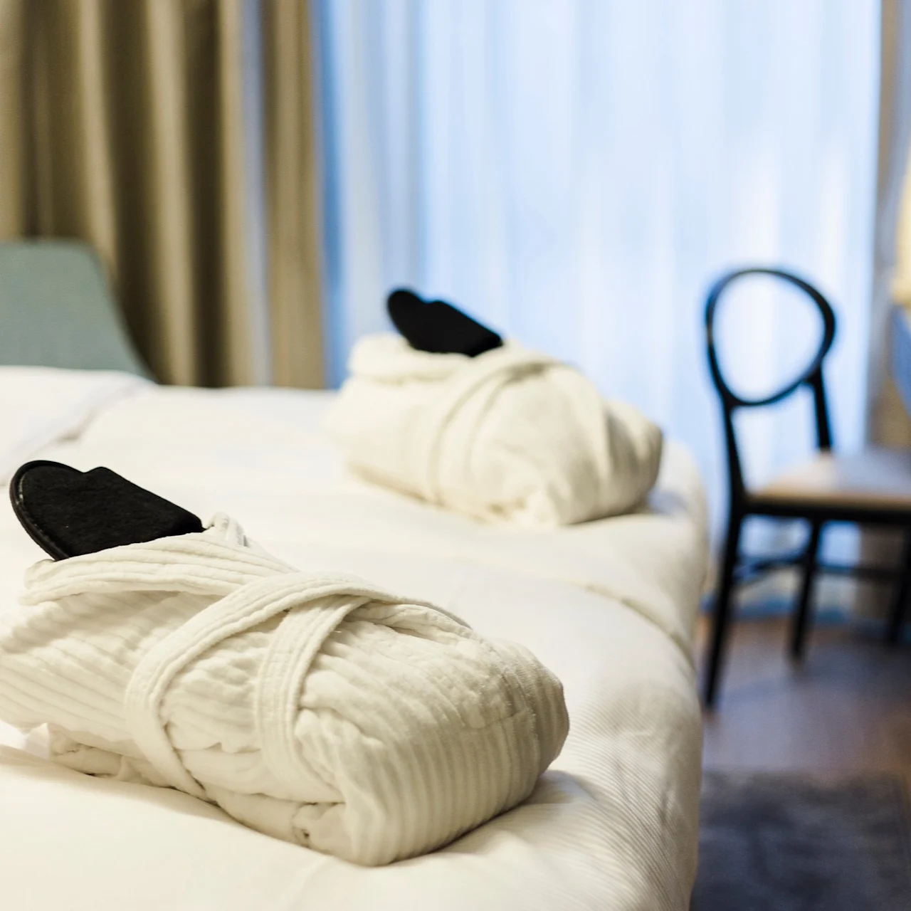 Bathrobes with slippers rest on a neatly made hotel bed; next to them, a chair and desk with a lamp create a minimalist, serene atmosphere.