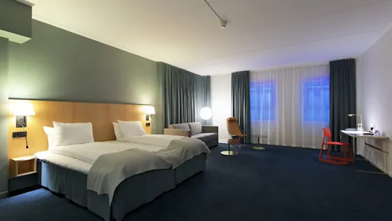 Spacious hotel room features two beds with white linens, a gray sofa, and a desk with a red chair. Large windows, blue curtains, and soft lighting enhance the modern interior.