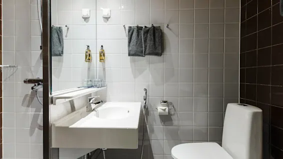 A white sink with hand soaps and a toilet are in a tiled bathroom. Gray towels hang above on hooks. Lighting reflects softly off the tiles. No text is present.