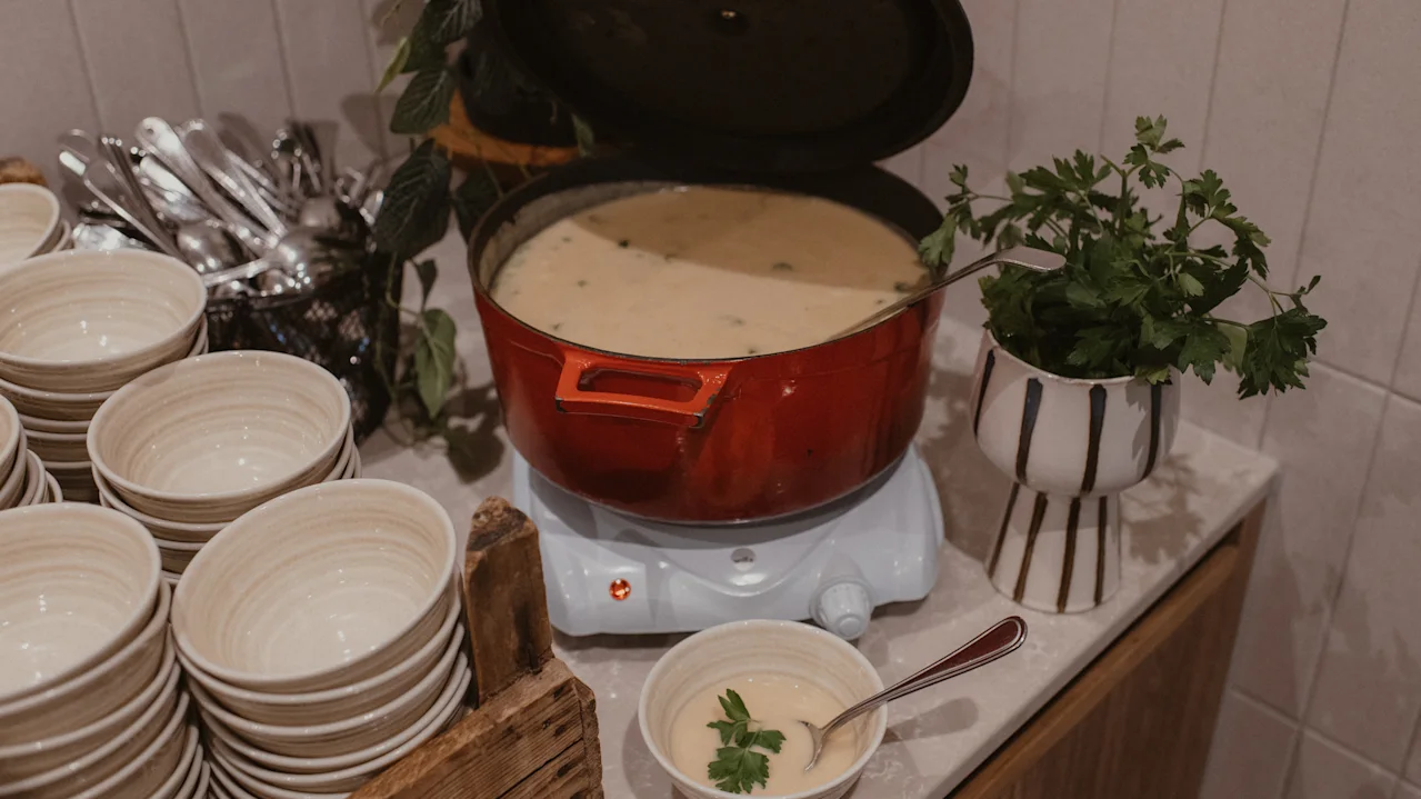 A red pot of creamy soup sits warming on a hot plate, surrounded by stacked bowls and fresh parsley in a planter on a counter.