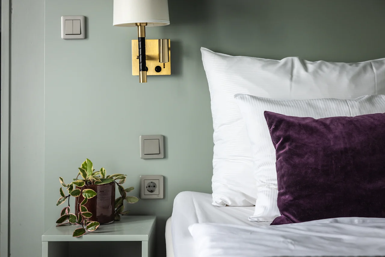 Kodikas makuuhuoneen maisema, jossa on siististi pedattu sänky purppuranvärisellä tyynyllä. Pieni kasvi on yöpöydällä ja seinävalo lisää lämpöä pehmeille vihreille seinille.