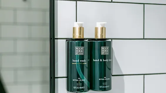 To mørkegrønne flasker med guldpumper mærket "hand wash" og "hand & body lotion", del af "The Ritual of Jing"-kollektionen, er monteret på en flisebelagt badeværelsesvæg.