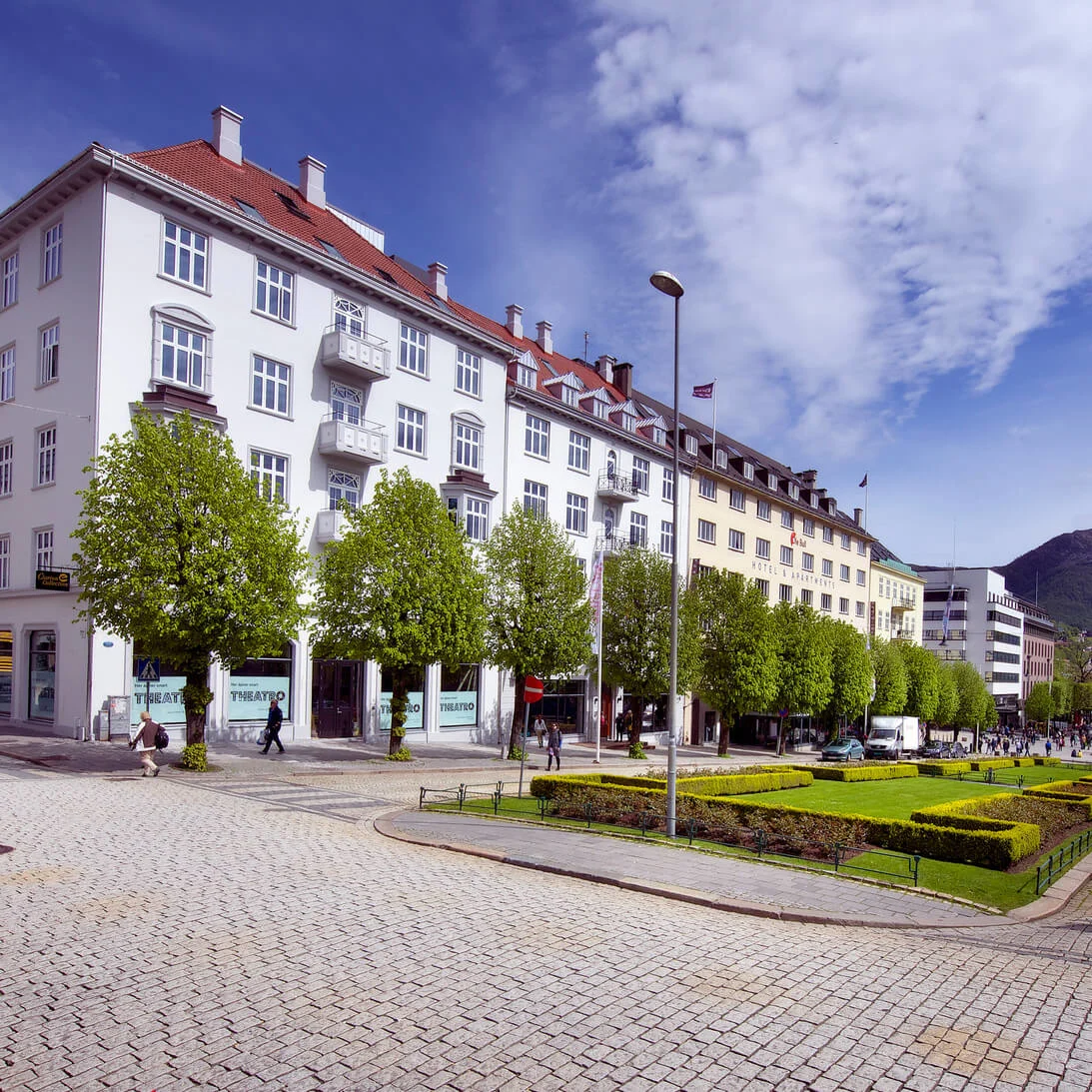 English: A tree-lined street borders a city park with geometric grass patterns; pedestrians walk on the cobblestone path, surrounding tall buildings bearing signage like "THEATRE."