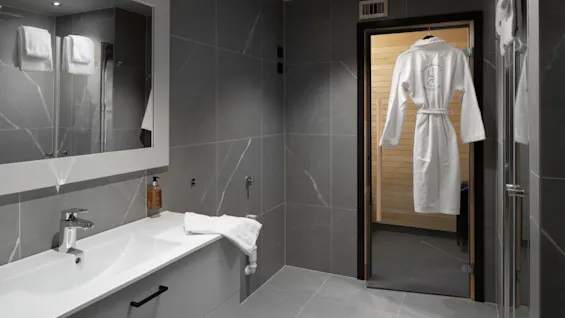 Et moderne badeværelse har en hvid vask og spejl, med en hængende badekåbe synlig gennem en dør, der fører til en sauna. Mørkegrå fliser dækker vægge og gulv.