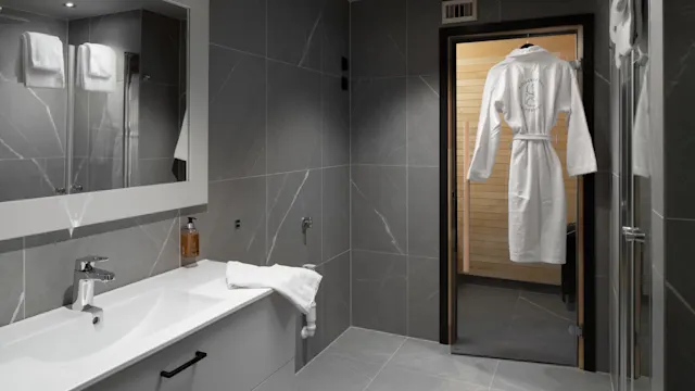A modern bathroom features a white sink and mirror, with a hanging robe visible through a door leading to a sauna. Dark gray tiles cover the walls and floor.
