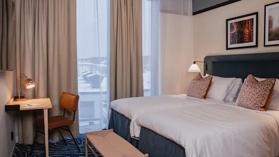 Ett prydligt hotellrum med en dubbelsäng med dekorativa kuddar, ett närliggande skrivbord med stol och lampa, och ett stort fönster med utsikt över en stadsmiljö.