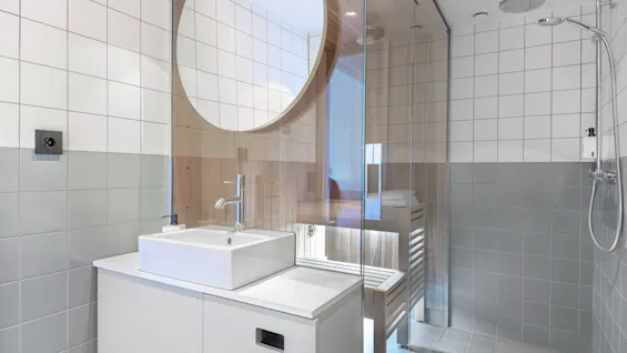 Moderne baderom har en firkantet vask med kran på en benk, ved siden av en dusj med glassvegger. Hvite og grå fliser dekker veggene, og forsterker det elegante designet.