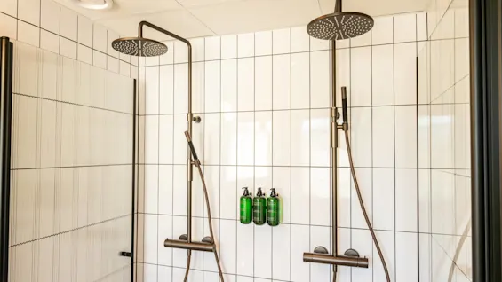 Two rainfall showerheads with handheld attachments are mounted on a white tiled wall in a modern bathroom. Three green bottles are attached to the wall between the showers.