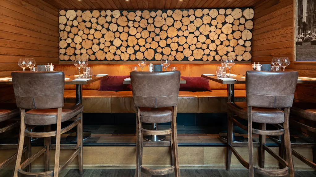 Spisebord er satt med glass og tallerkener, arrangert rundt skinnstoler og benker. En vegg med tredesign av tømmerstokker dekorerer bakgrunnen i en varmt opplyst restaurant.