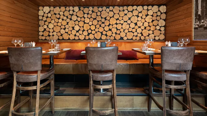 Dining tables are set with glasses and plates, arranged around leather chairs and benches. A wooden log wall design decorates the background in a warmly lit restaurant interior.