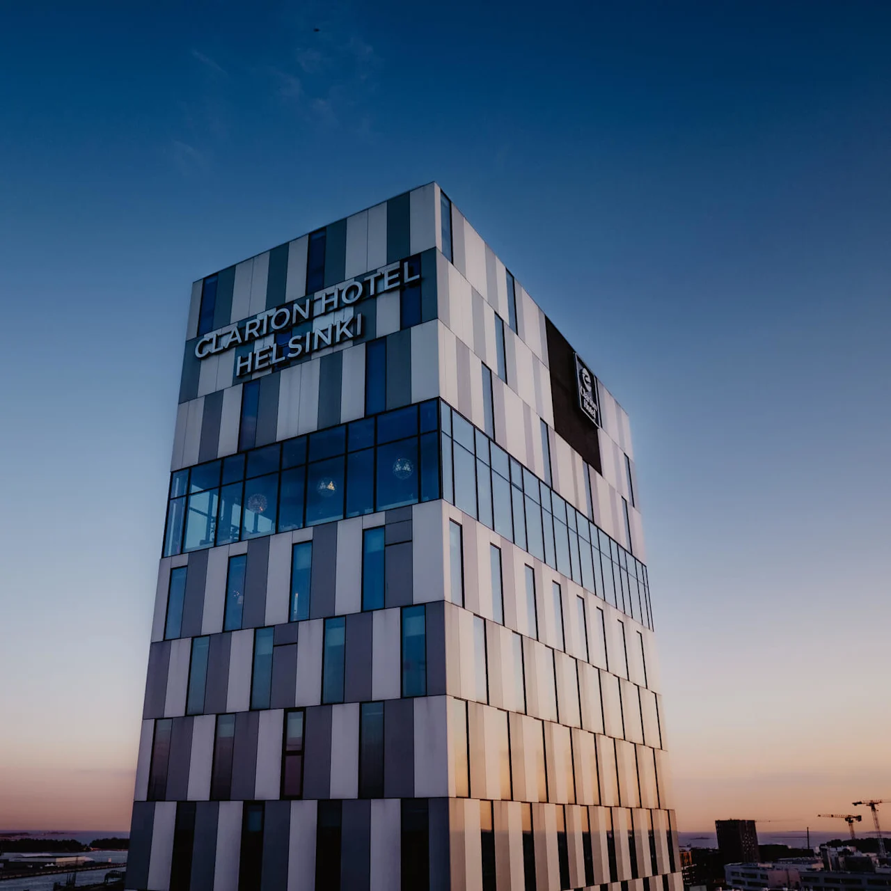 Et moderne højhus, kaldet "CLARION HOTEL HELSINKI," står mod en skumringshimmel med bybillede og hav i baggrunden.