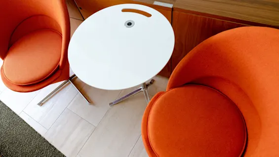 Två orange stolar omger ett litet vitt bord med ett handtag. Ett träskåp märkt "Glasses inside" finns i bakgrunden på ett kaklat golv.