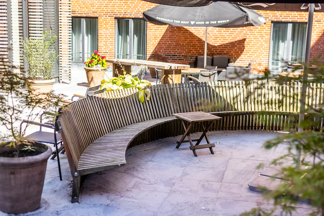 An inviting outdoor patio at Comfort Hotel Vesterbro with a curved wooden bench, small table, and umbrella that reads PROBABLY THE BEST BEER IN THE WORLD.