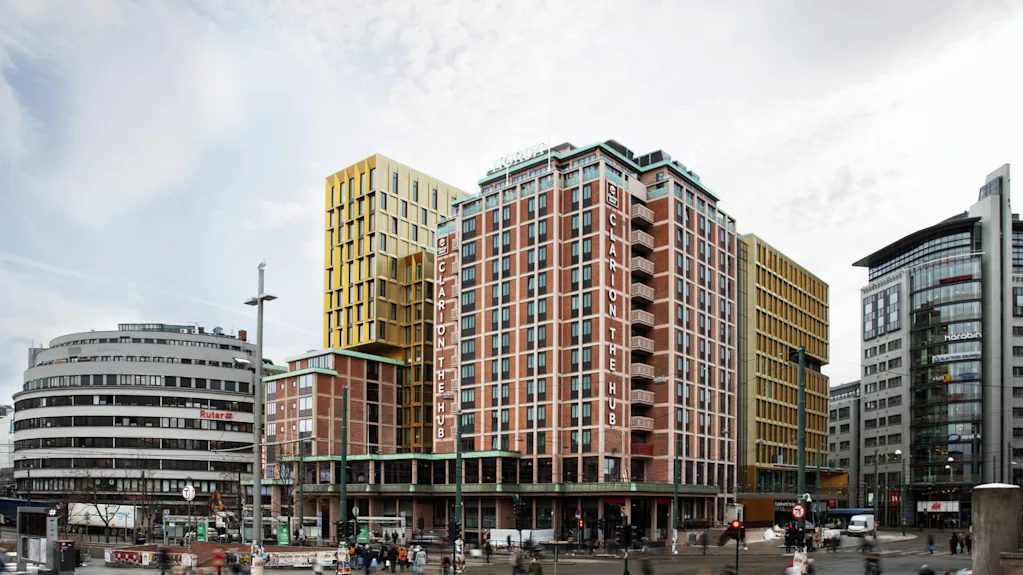 Clarion The Hub building stands tall, displaying "CLARION THE HUB" on its facade in an urban setting. Surrounding it are modern and rounded office buildings. People and vehicles are visible on the street. 