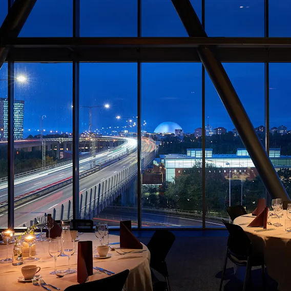 Round tables set with elegant dining ware sit by large windows, overlooking a highway with light trails from cars, and a skyline with a distinctive dome.