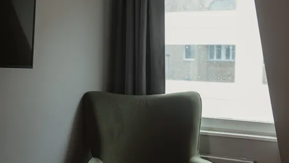 A green armchair sits by a large window with gray curtains, in a minimalist room with light grey walls and part of a bed visible.