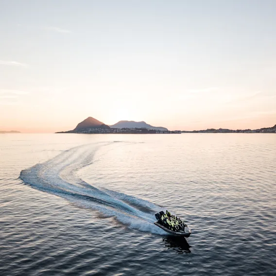 Luftfoto av en rask RIB-båt som lager en svingete hekkbølge i det blikkstille fjordvannet i solnedgang.