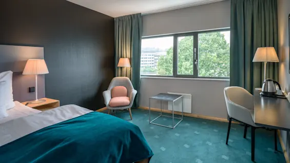 Et ryddeligt hotelværelse har en seng med turkis sengetøj, et skrivebord med en elkedel og en lænestol ved et vindue med udsigt over træer, hvilket skaber en hyggelig, moderne atmosfære.