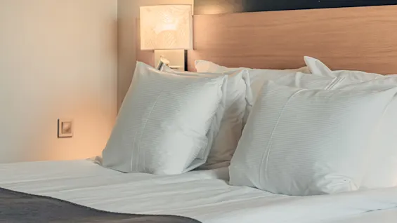 White pillows rest neatly against a wooden headboard, with a textured lamp emitting soft light beside them on a smooth, neutral-toned wall.