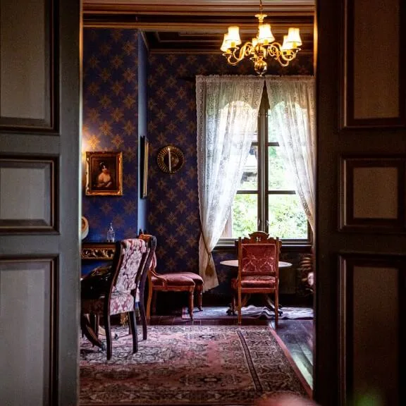 A vintage room features ornate chairs around a table, a chandelier above, blue-patterned wallpaper, and lace curtains framing a window, viewed through a wooden doorway.